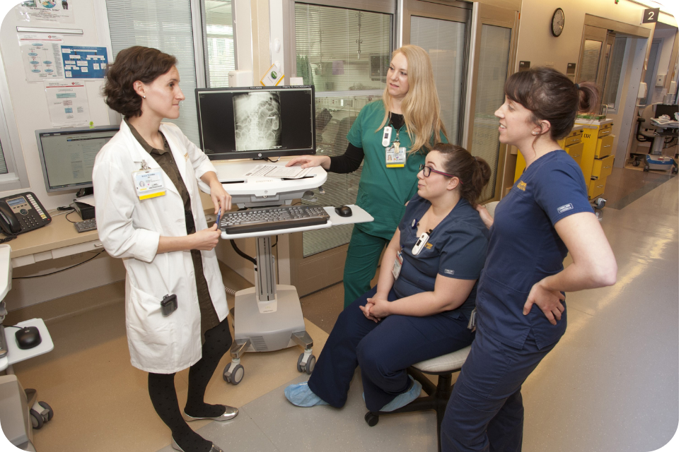 Doctor in white coat explains monitor scan to three nurses in scrubs in hospital room