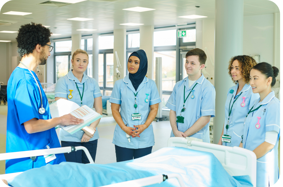 Diverse nurses in light-blue scrubs listening to instructor beside hospital bed