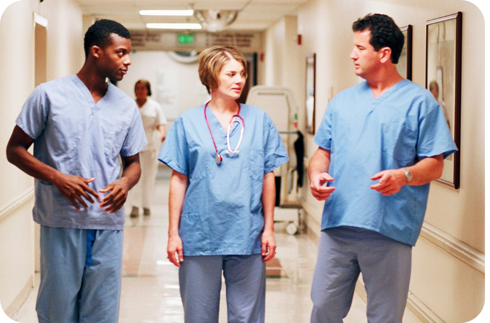 Three healthcare workers in blue scrubs walking and talking in a hospital corridor