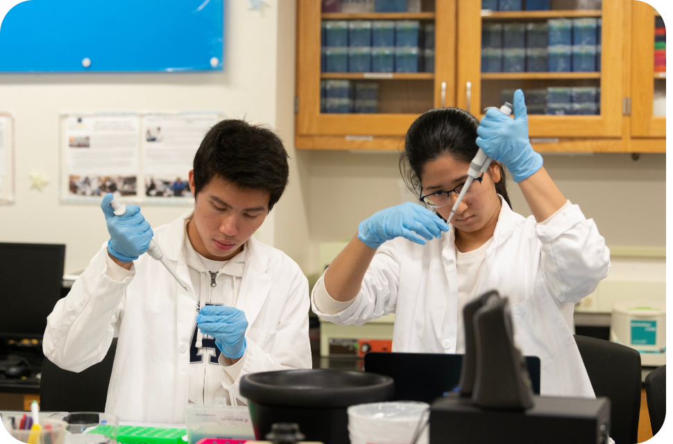 Two researchers in white lab coats using pipettes at a lab bench.