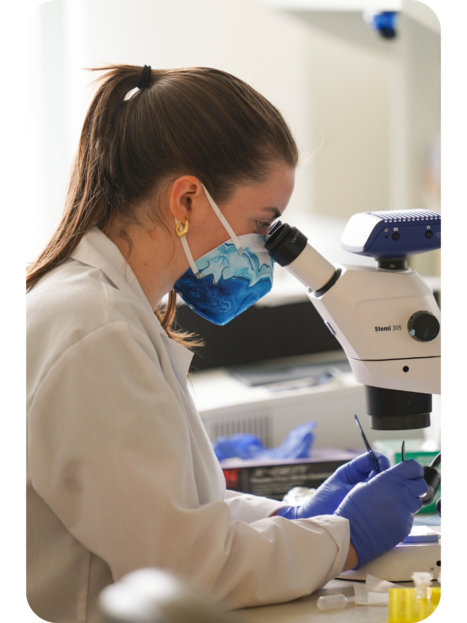 Female scientist in lab coat, blue mask and gloves, peering into microscope