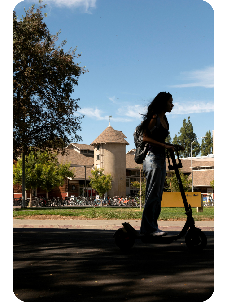 Silhouetted person riding an electric scooter past trees and a round-towered building