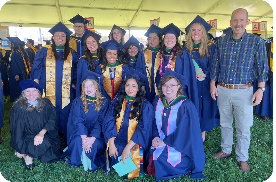 Group of smiling graduates in navy caps and gowns with stoles and leis under a tent