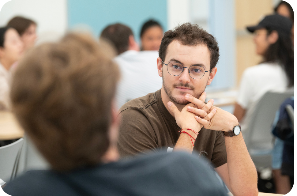 Young man with glasses and brown shirt attentively listening in a classroom, blurred students behind
