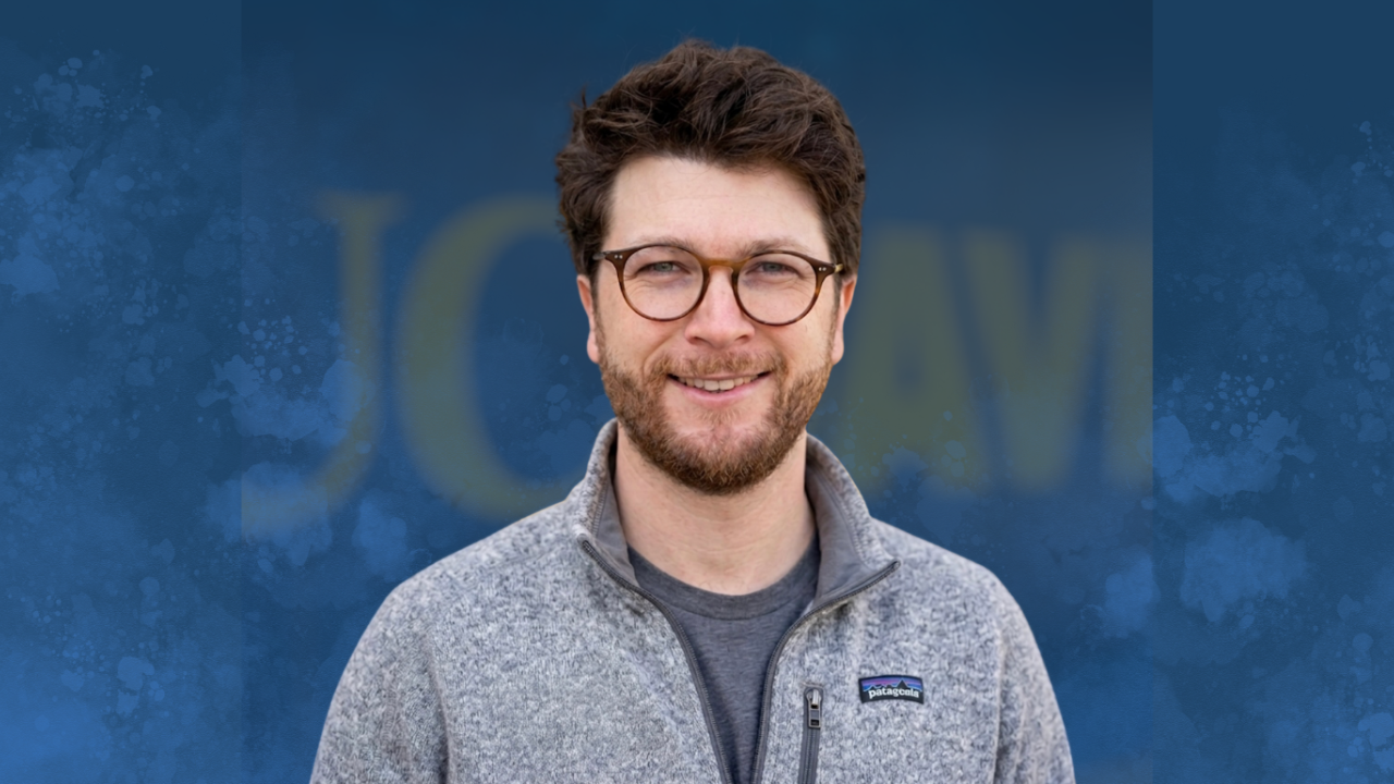 Headshot photograph of smiling young man with glasses and curly hair on blue textured backdrop