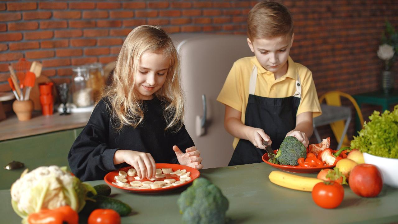 Two children in aprons preparing vegetables on a kitchen counter with brick wall background