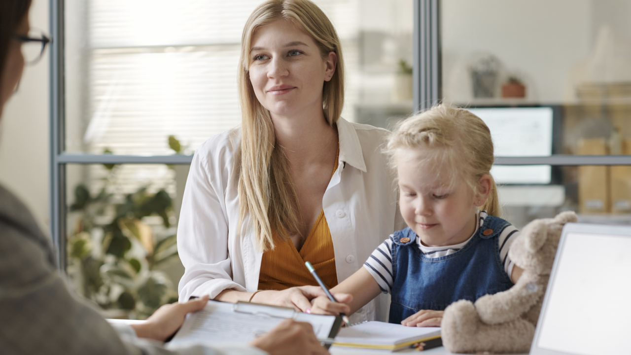 Mother smiling beside daughter who colors with teddy at an office desk