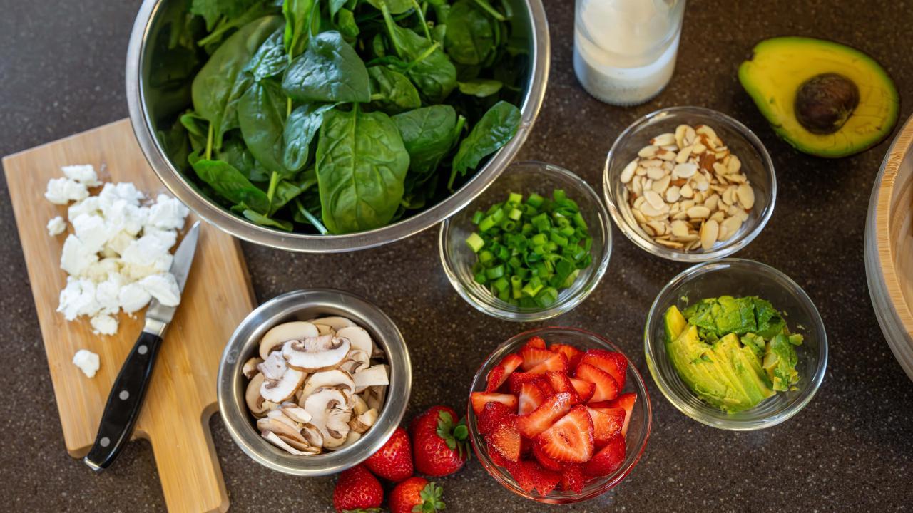 A kitchen counter with an array of ingredients including spinach, strawberries, avocado, almonds, cheese and mushrooms to prepare a salad.