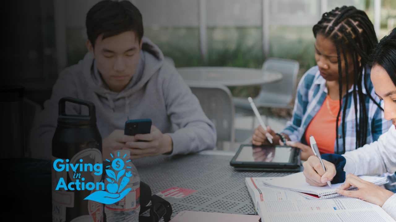 Three students studying at a table outdoors; woman writing; small blue logo left (photo)