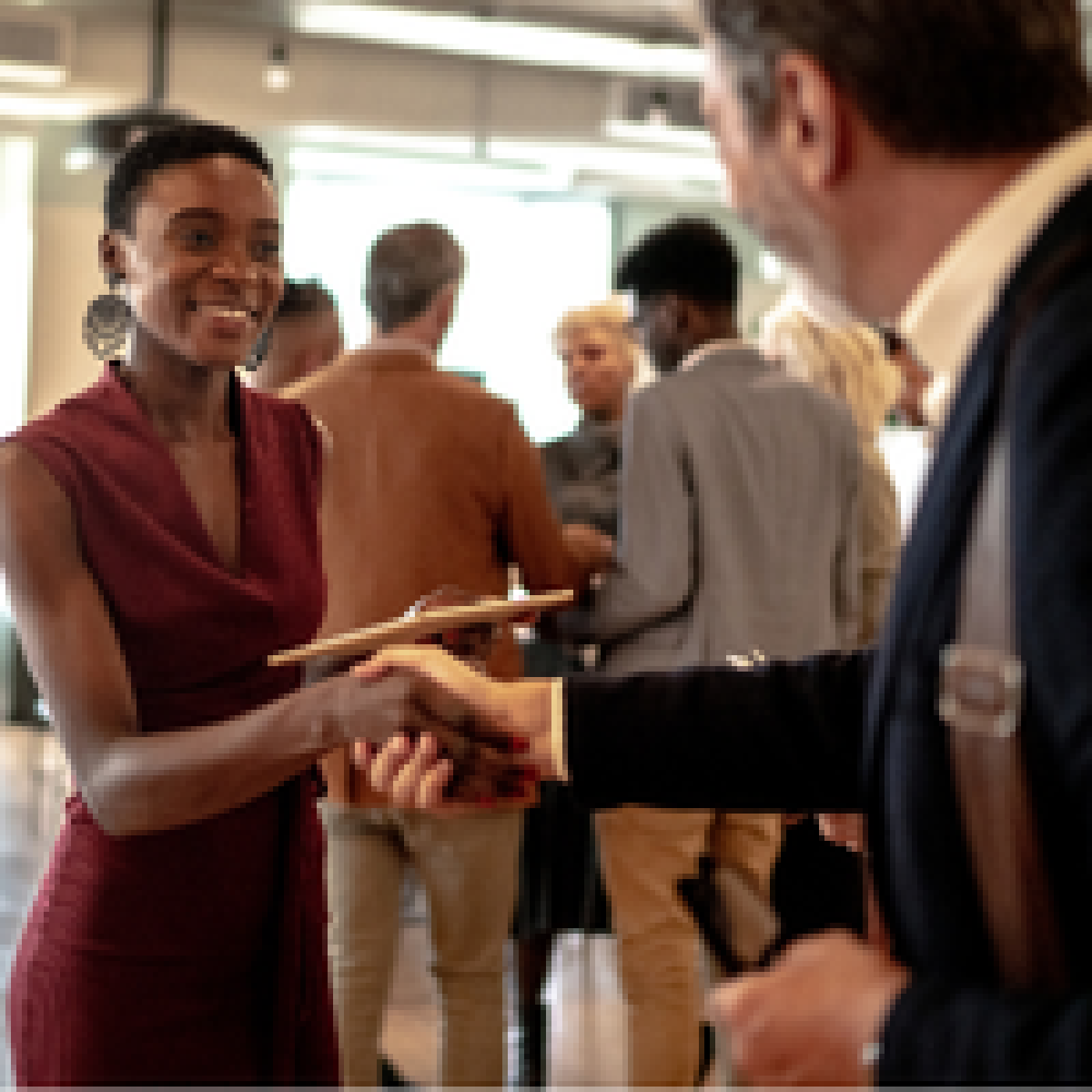 Smiling woman in maroon dress shakes hands while offering a notepad at a networking event