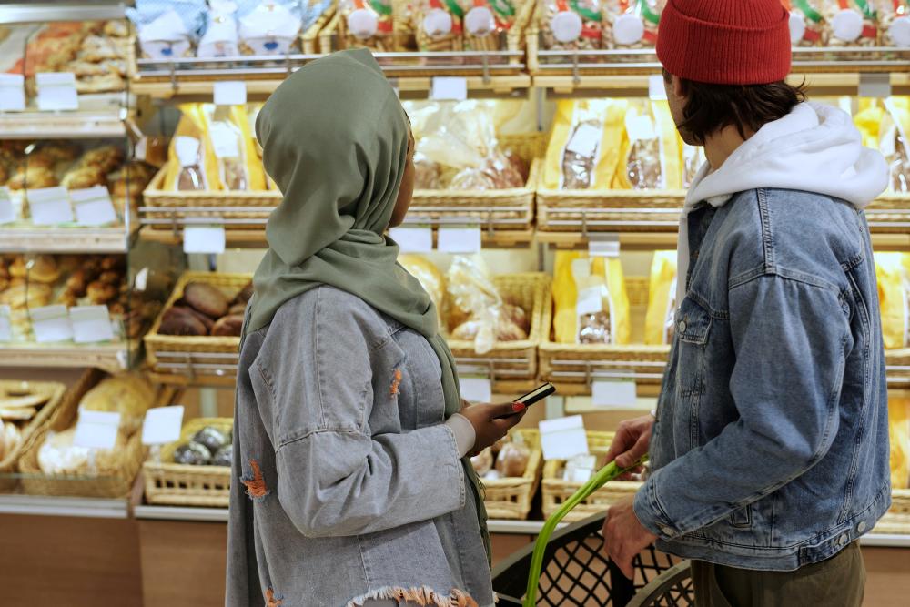 Two women selecting bread in a bakery, one wearing a green hijab and one a red beanie
