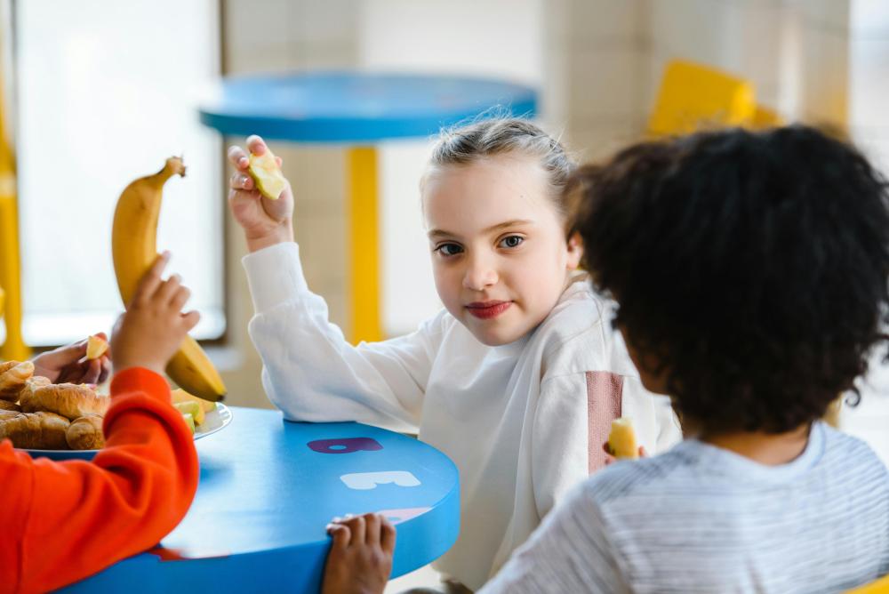 Girl in white hoodie peeling a banana at a blue classroom table with classmates