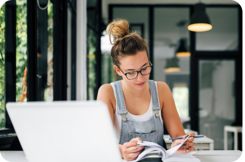 Young woman in glasses and denim overalls studying with laptop, phone, and notebook