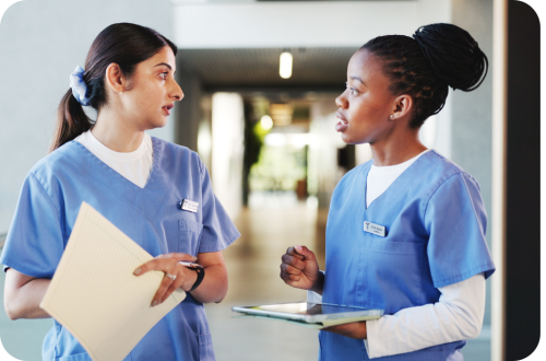 Two nurses in blue scrubs talking in a hospital corridor, holding charts (photo)