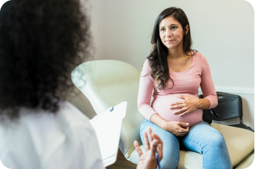 Pregnant woman seated with hands on belly, speaking with clinician in exam room