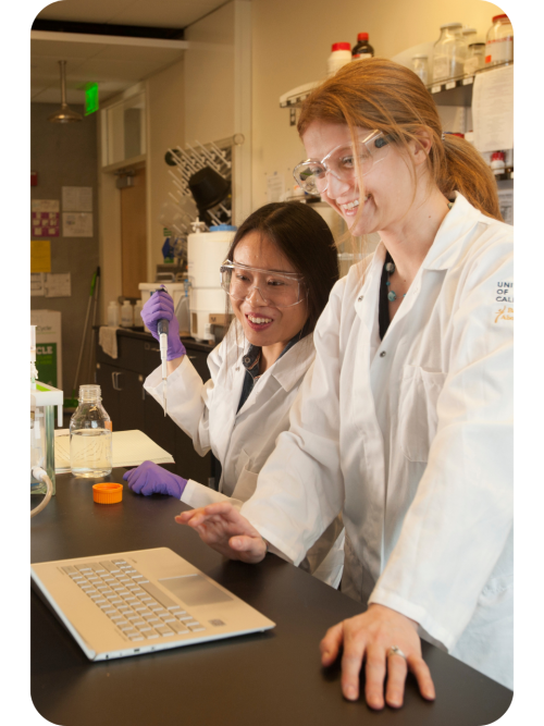 Two scientists in lab coats and gloves smiling at a laptop in a bright lab (photo)