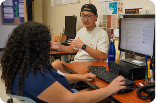 Man in backward cap gesturing to woman with curly hair at a desk with computer monitors.