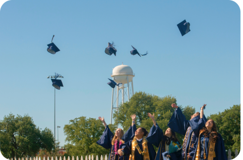 Graduates in navy gowns toss mortarboards into blue sky near a water tower