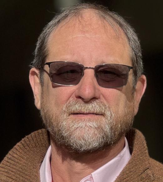 Headshot photo of older man with gray beard and sunglasses, neutral expression, brown jacket