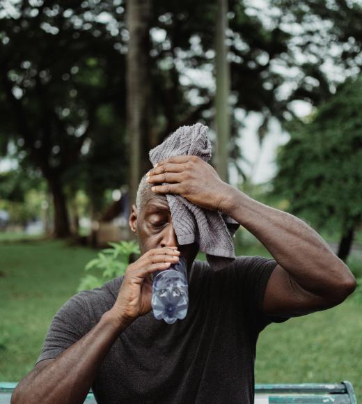 Man on park bench covering face with gray towel, outdoor photo.
