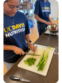 Two people in UC Davis Nutrition shirts chopping green herbs and stirring a pan