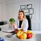 Smiling woman in white coat at office desk holding broccoli; bowl of fruit, certificates behind