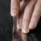 Fingertips guiding a scribing blade along a metal ruler to score a dark sheet