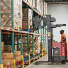 Warehouse worker in orange hard hat operating red pallet stacker between tall shelves of boxes