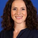 Professional headshot of smiling woman with curly brown hair against blue background