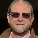 Headshot photo of older man with gray beard and sunglasses, neutral expression, brown jacket