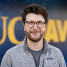 Headshot photo of a smiling bearded man with glasses against a blurred blue-yellow background