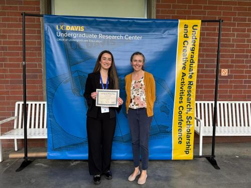 Two smiling women, one holding a certificate, standing before a UC Davis blue-and-gold banner